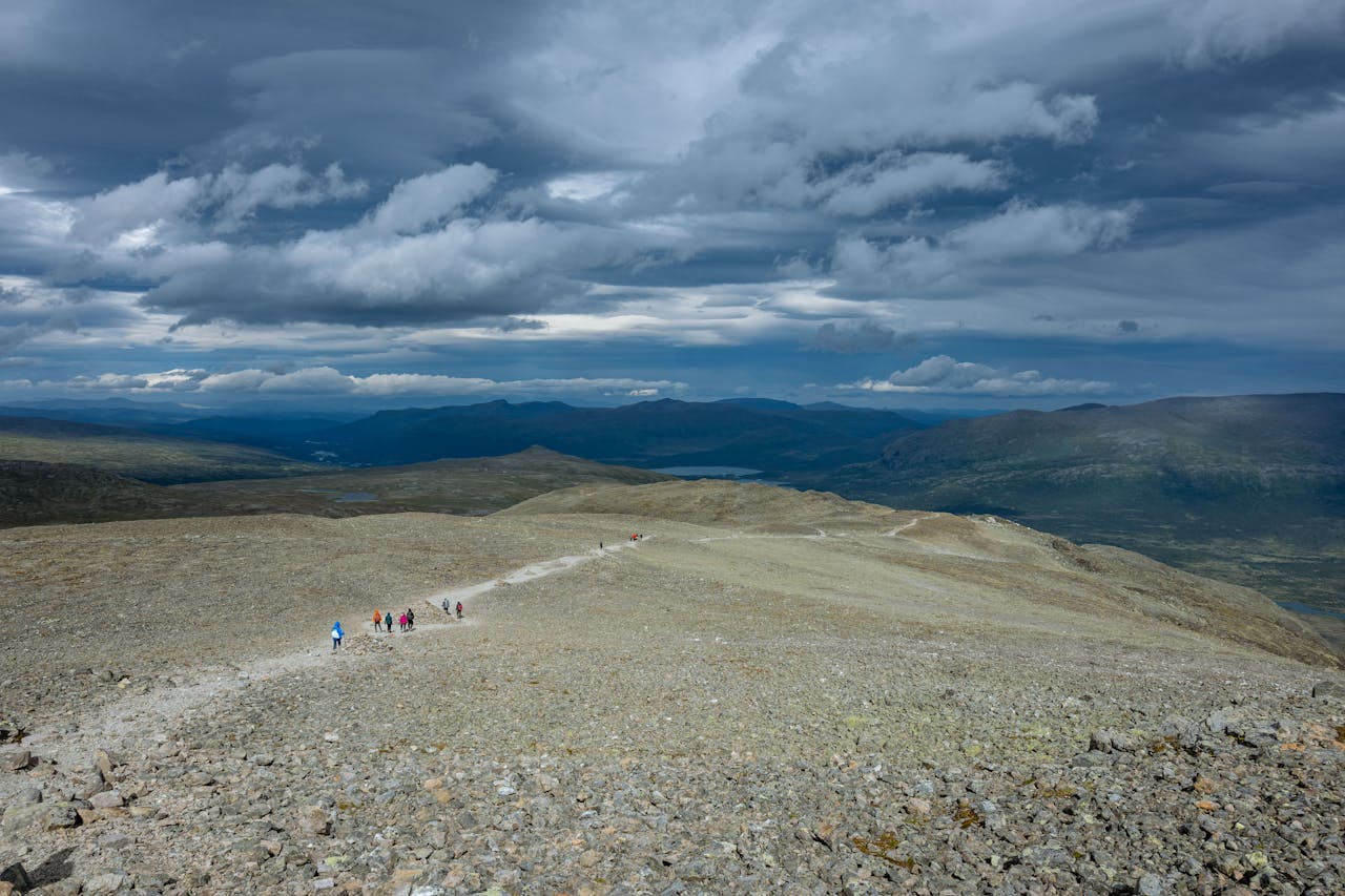 heros-img A group of hikers on a rocky mountain trail in Innlandet, Norway with dramatic clouds above.