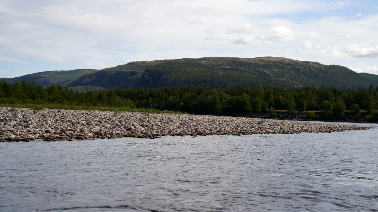 Idyllic river view with pebble shore, forested hills, and wide sky background.
